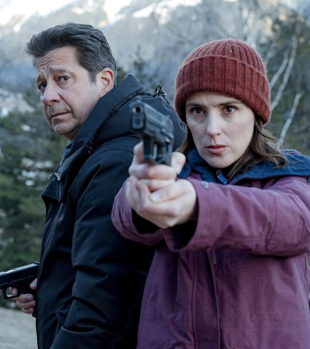 Andréas (Laurent Gerra) und Constance (Clémentine Poidatz) stehen tagsüber in einer winterlichen Berglandschaft. Constance richtet eine Waffe seitlich nach vorne. Neben ihr blickt Andréas ebenfalls angespannt nach vorne.