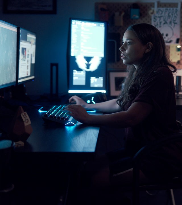 Halbtotale: Kelly (Jessica Parker Kennedy) sitzt in einem dunklen Zimmer an einem Schreibtisch mit mehreren Bildschirmen.