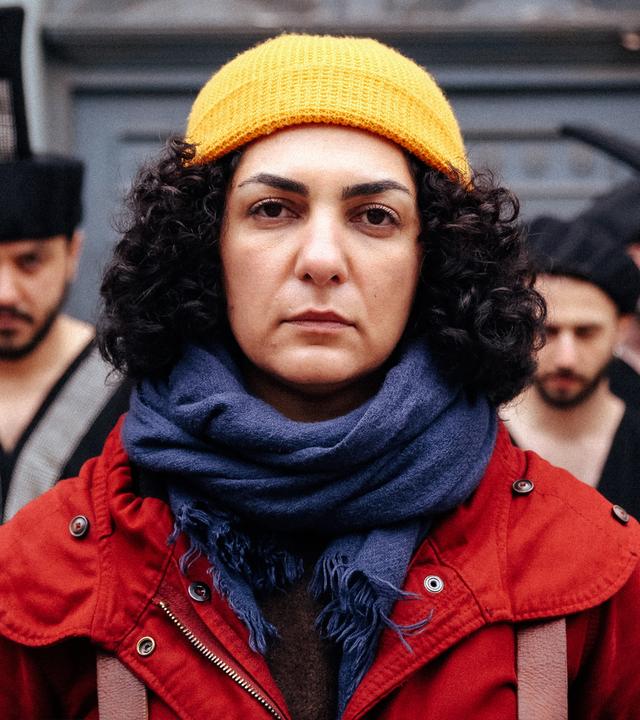 Narges (Baharak Abdolifard) blickt nach vorn. Sie trägt eine rote Jacke, einen blauen Schal und eine gelbe Mütze. Hinter ihr stehen Männer in schwarzen Kleidern und mit schwarzen Hüten.
