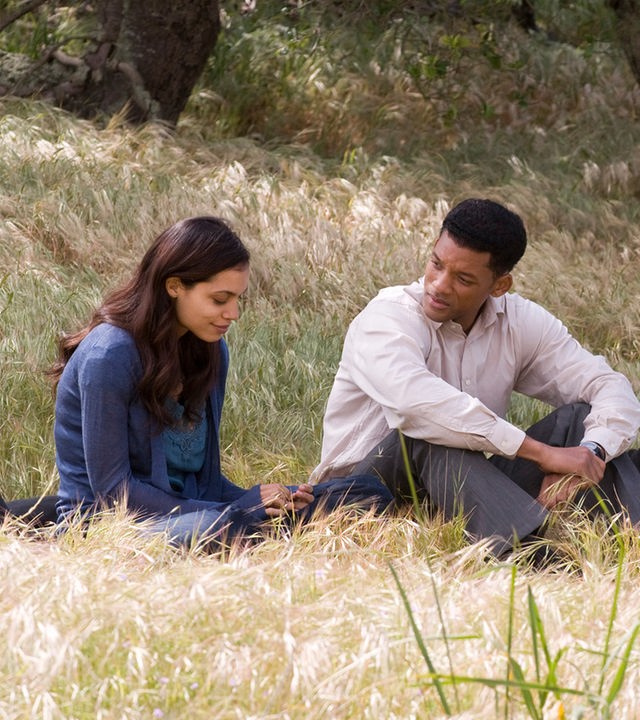 "Sieben Leben": Ben (Will Smith), Emily (Rosario Dawson) und ein schwarz-weißer Hund sitzen im hohen Gras. Während Emily nach unten blickt, wendet Ben sich ihr zu. Der Hund schaut aufmerksam in eine andere Richtung.