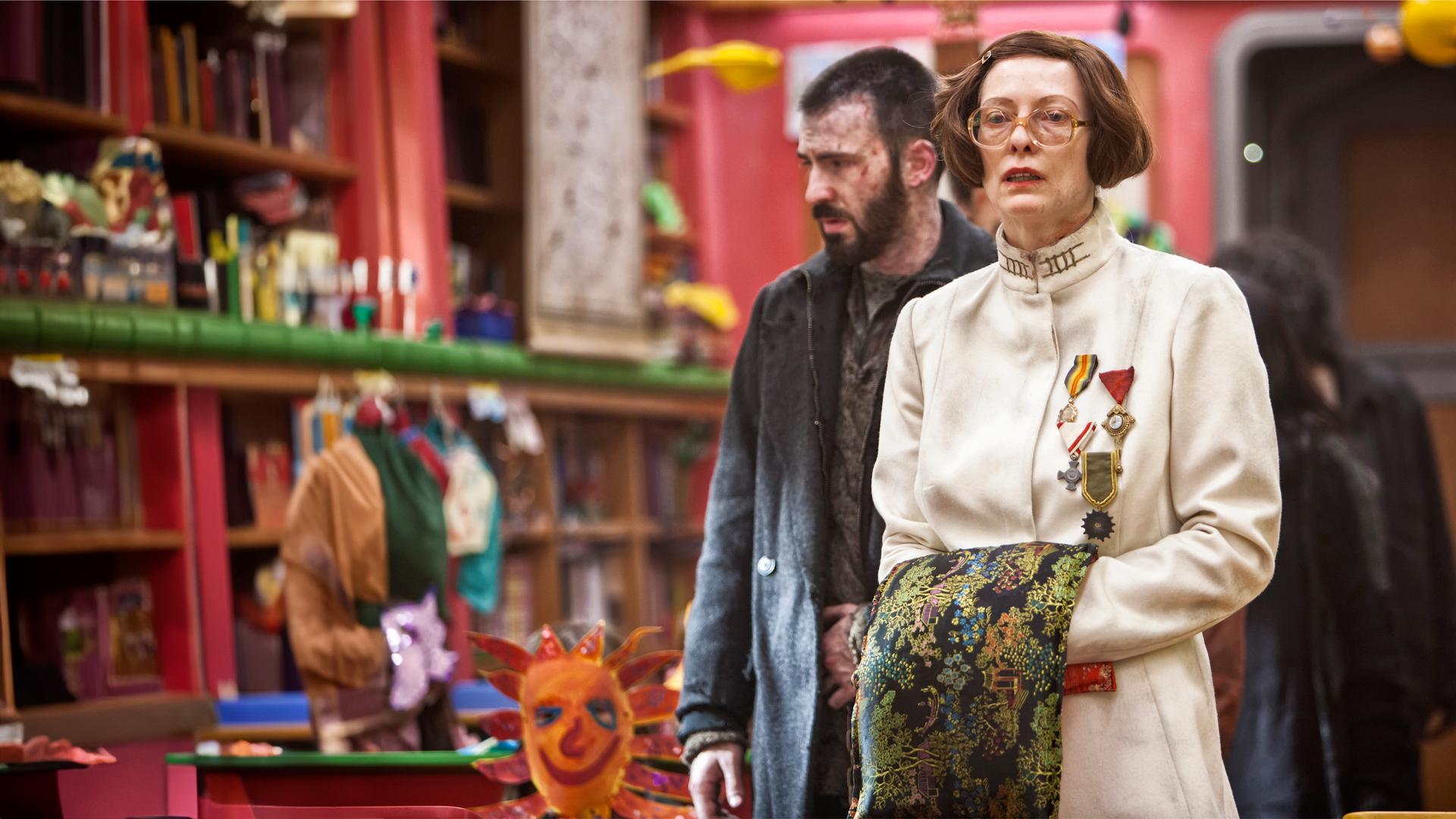 Mason (Tilda Swinton), mit großer Brille und pagenartiger Frisur, trägt einen weißen Mantel mit Stehkragen. Sie hat sich mehrere Orden angeheftet. Ihre Hände sind vor dem Bauch zusammengeführt und mit einem verzierten Tuch verdeckt. Sie schaut verwirrt. Leicht seitlich hinter ihr steht Curtis (Chris Evans) in heruntergekommener Kleidung. Er ist etwas unscharf abgebildet. Im Hintergrund sind ebenfalls unscharf ein Kinderklassenzimmer und selbstgebastelte Papiermasken zu erkennen.