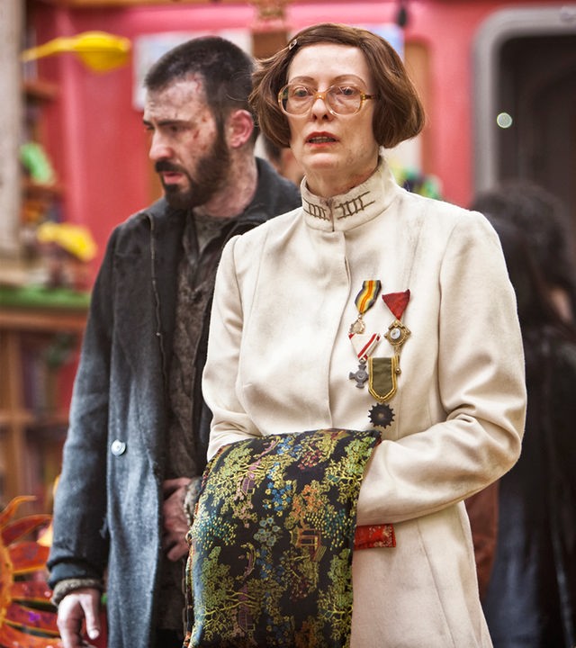 Mason (Tilda Swinton), mit großer Brille und pagenartiger Frisur, trägt einen weißen Mantel mit Stehkragen. Sie hat sich mehrere Orden angeheftet. Ihre Hände sind vor dem Bauch zusammengeführt und mit einem verzierten Tuch verdeckt. Sie schaut verwirrt. Leicht seitlich hinter ihr steht Curtis (Chris Evans) in heruntergekommener Kleidung. Er ist etwas unscharf abgebildet. Im Hintergrund sind ebenfalls unscharf ein Kinderklassenzimmer und selbstgebastelte Papiermasken zu erkennen.