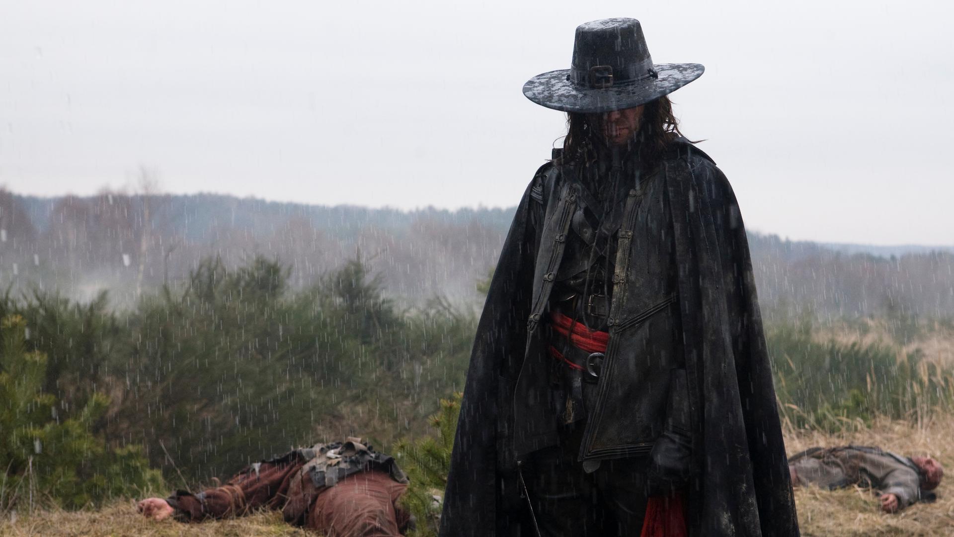 "Solomon Kane": Solomon Kane (James Purefoy) steht auf einer Wiese im strömenden Regen. Hinter ihm liegen zwei tote Männer (Komparsen) im von der Sonne ausgeblichenen Gras.