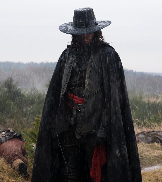 "Solomon Kane": Solomon Kane (James Purefoy) steht auf einer Wiese im strömenden Regen. Hinter ihm liegen zwei tote Männer (Komparsen) im von der Sonne ausgeblichenen Gras.