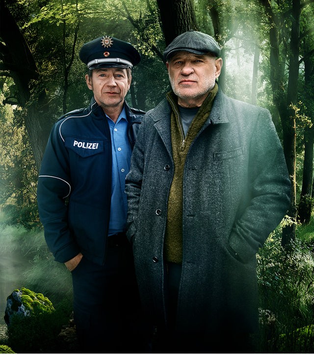 Thorsten Krüger (Christian Redl, links.) und Martin Fichte (Thorsten Merten, rechts) stehen in einer Waldlanschaft. Krüger trägt einen grauen Mantel, einen olivgrünen Strickpulover und eine graue Schiebermütze. Seine Hände hat er in der Manteltasche. Fichte trägt Polizeiuniform und hat seine Hände in der Hosentasche. Im Hintergrund ist ein kleiner Bach zu sehen, der durch einen Wald fließt. rechts und links des Baches ragen Bäume empor. Die gnaze Szenerie ist geprägt von Nebelschwaden und derdurch den Nebel  durchscheinenden Sonne.