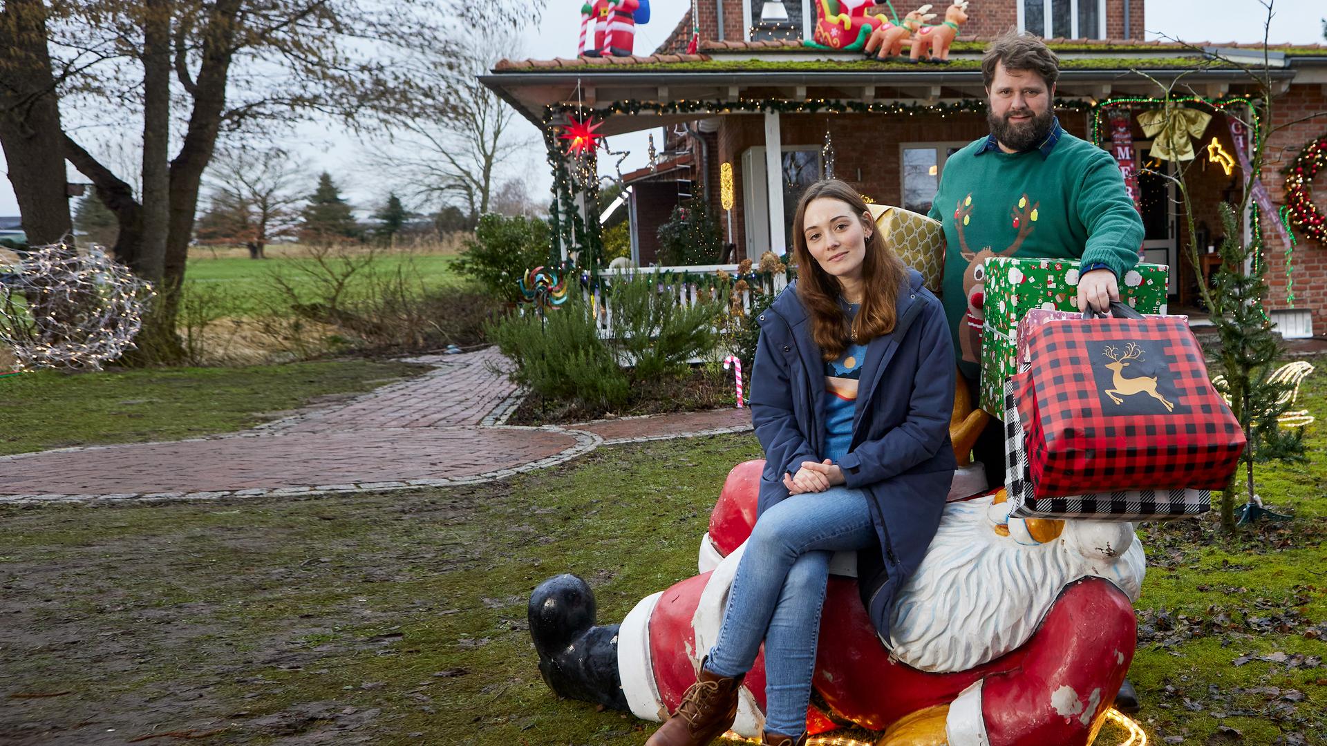 Liane (Maria Ehrich) sitzt auf einer liegenden Weihnachtsmann-Dekoration auf dem Rasen vor einem weihnachtlich geschmückten Haus. Sie schaut amüsiert und blickt in die Kamera. Hinter ihr steht Hinnerk (Antoine Monot), der viele große, bunte Weihachtsgeschenke unter den Armen hält Auch er schaut lachend in die Kamera.