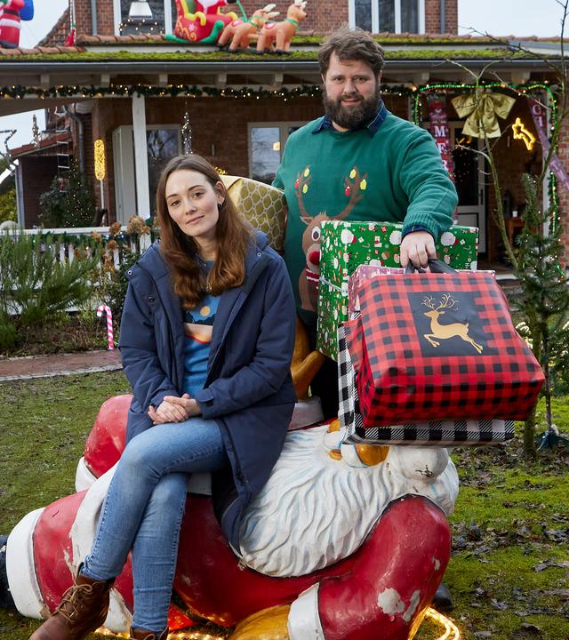 Liane (Maria Ehrich) sitzt auf einer liegenden Weihnachtsmann-Dekoration auf dem Rasen vor einem weihnachtlich geschmückten Haus. Sie schaut amüsiert und blickt in die Kamera. Hinter ihr steht Hinnerk (Antoine Monot), der viele große, bunte Weihachtsgeschenke unter den Armen hält Auch er schaut lachend in die Kamera.
