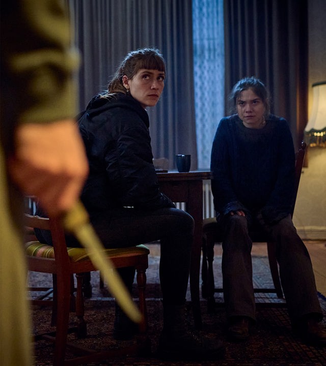 „STRALSUND – Dunkle Tiefen“: Jule (Sophie Pfennigstorf) und Ella (Hannah Schiller) sitzen im Wohnzimmer an einem Tisch. Beide Frauen drehen sich angespannt zu einem Unbekannten um, der im Vordergrund steht und ein Messer in seiner rechten Hand hält.