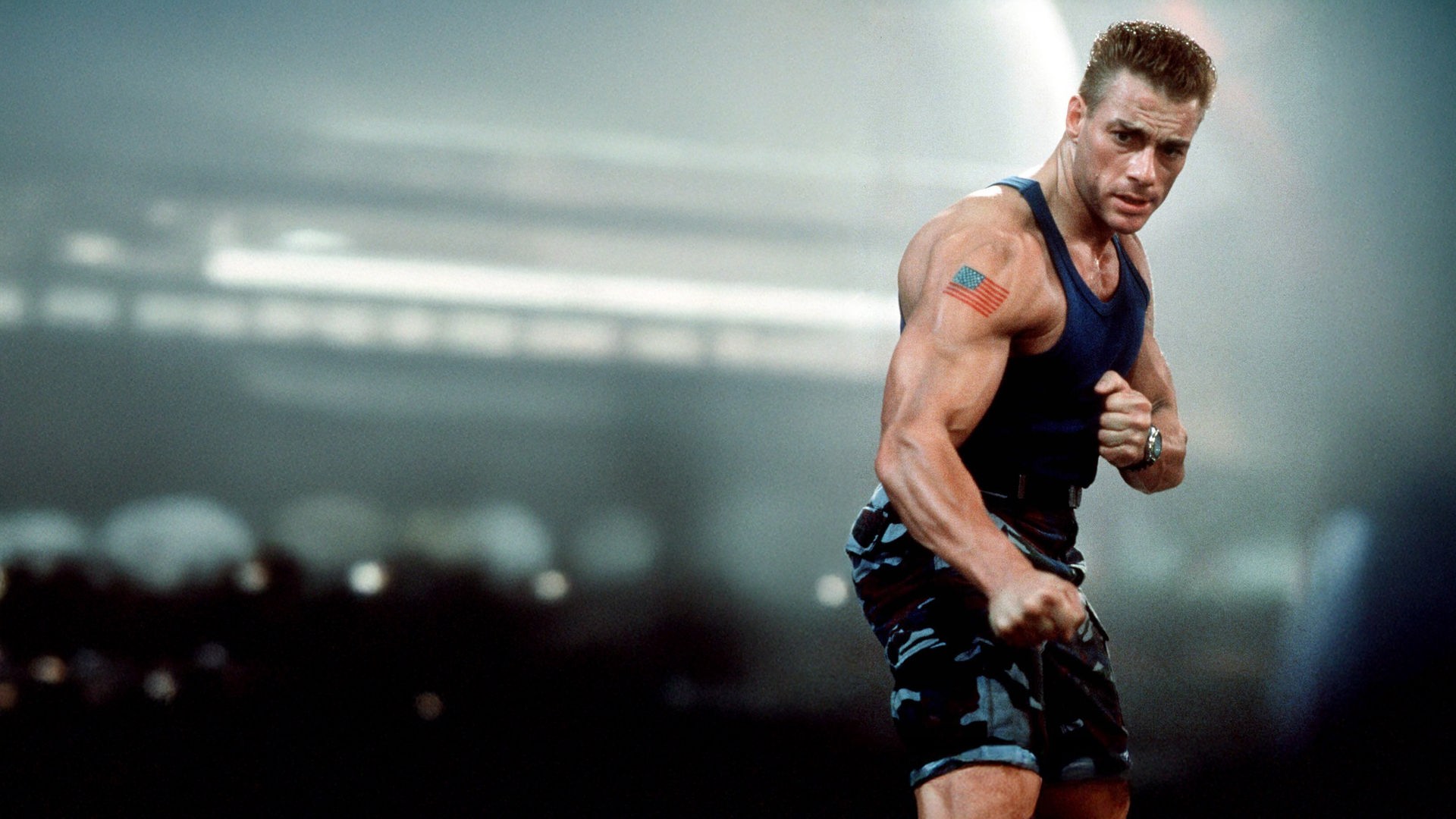 „Street Fighter - Die entscheidende Schlacht“ - Colonel William F. Guile (Jean-Claude Van Damme) steht in kampfbereiter Pose mit geballten Fäusten vor einem verschwommenen Hintergrund. Der muskulöse Charakter trägt ein blaues Unterhemd, eine Tarnhose sowie ein Tattoo der US-Flagge auf seinem rechten Oberarm.