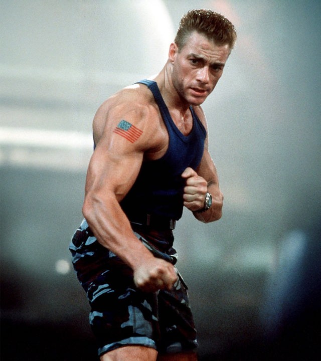 „Street Fighter - Die entscheidende Schlacht“ - Colonel William F. Guile (Jean-Claude Van Damme) steht in kampfbereiter Pose mit geballten Fäusten vor einem verschwommenen Hintergrund. Der muskulöse Charakter trägt ein blaues Unterhemd, eine Tarnhose sowie ein Tattoo der US-Flagge auf seinem rechten Oberarm.