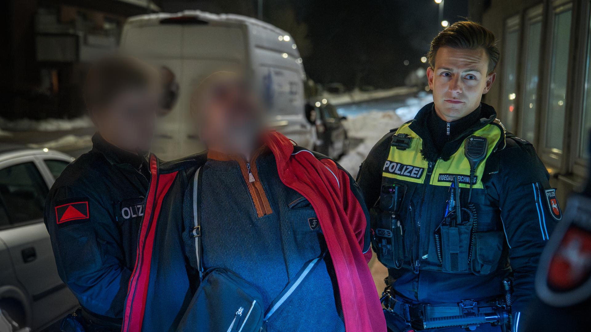 Ein betrunkener Mann in roter Jacke wird von zwei Polizisten in Gewahrsam genommen.
