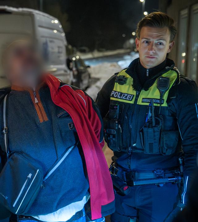 Ein betrunkener Mann in roter Jacke wird von zwei Polizisten in Gewahrsam genommen.