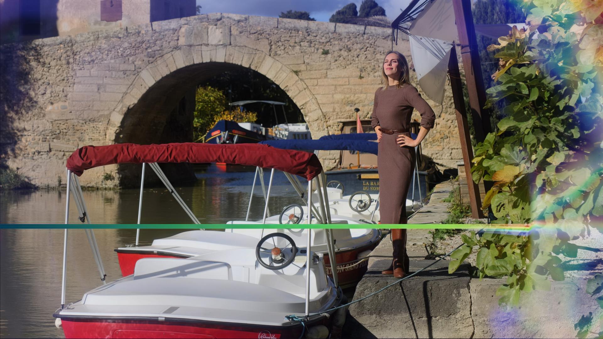 Die Auswanderin Jelena Weber steht neben einem Fluss, auf dem 3 Boote zu Wasser gelassen sind und über den im Hintergund eine bogenförmige Brücke führt.