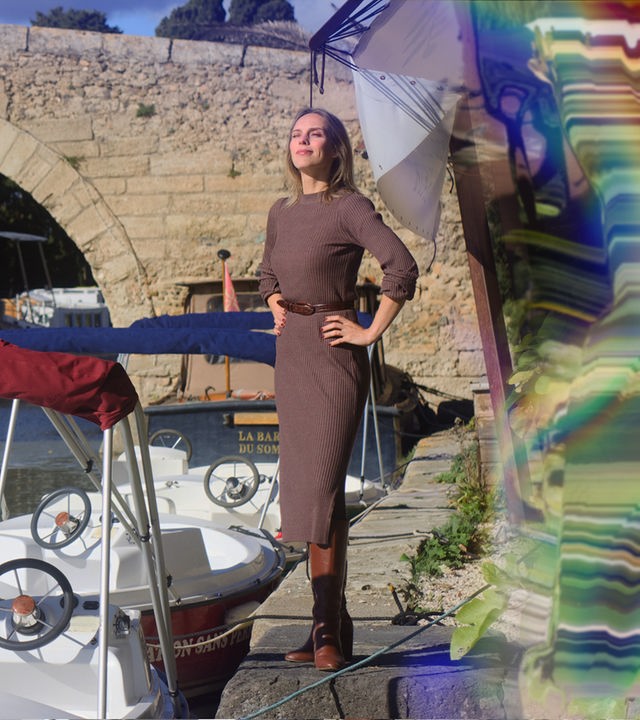 Die Auswanderin Jelena Weber steht neben einem Fluss, auf dem 3 Boote zu Wasser gelassen sind und über den im Hintergund eine bogenförmige Brücke führt.