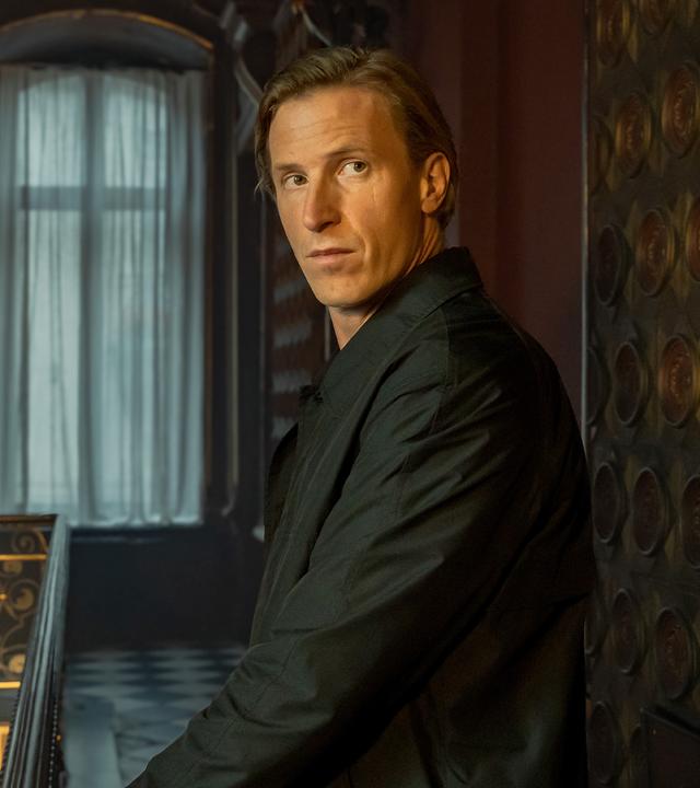 Carl Winterscheid (Golo Euler) steht in einem feudalen Treppenhaus auf einer Galerie und hat sich umgedreht. Eine Hand hat er auf dem gusseisernen Geländer liegen. Im Hintergrund der dunkel wirkenden Galerie sind zwei große Fenster mit hellen Vorhängen.