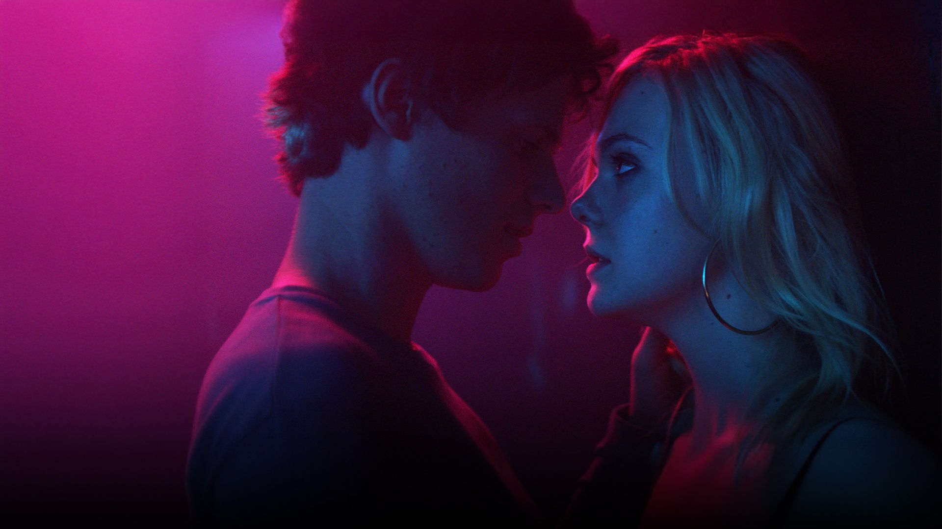 Teen Spirit": Nahaufnahme - Keyan Spears (Ruairi O'Connor) beugt sich mit intensivem Augenkontakt zu der nah vor ihm an einer Wand lehnenden Violet (Elle Fanning) zu einem Kuss vor. Die beiden, die im Profil zu sehen sind, werden von pinkfarbenem Licht angestrahlt.
