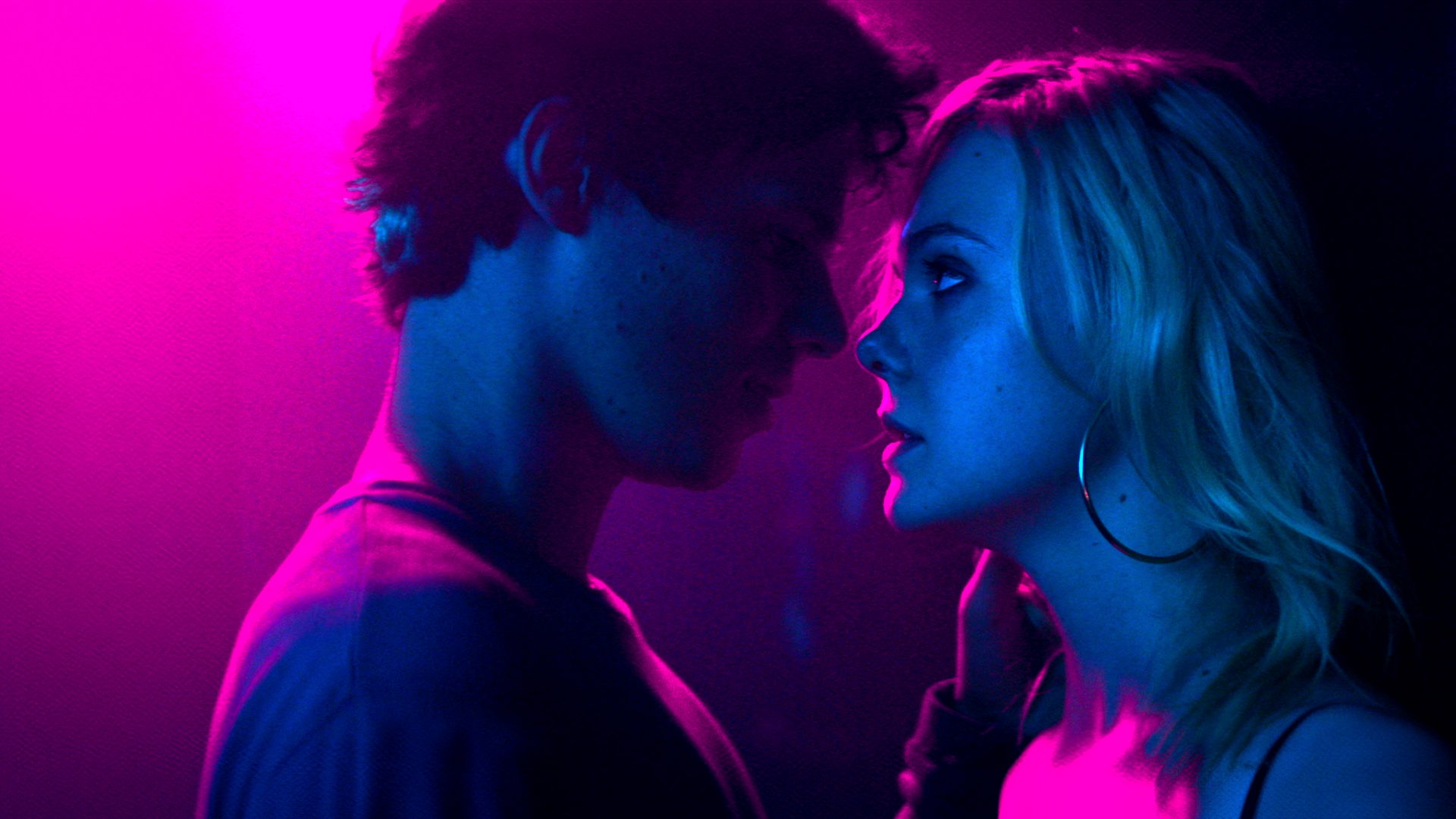 Teen Spirit": Nahaufnahme - Keyan Spears (Ruairi O'Connor) beugt sich mit intensivem Augenkontakt zu der nah vor ihm an einer Wand lehnenden Violet (Elle Fanning) zu einem Kuss vor. Die beiden, die im Profil zu sehen sind, werden von pinkfarbenem Licht angestrahlt.