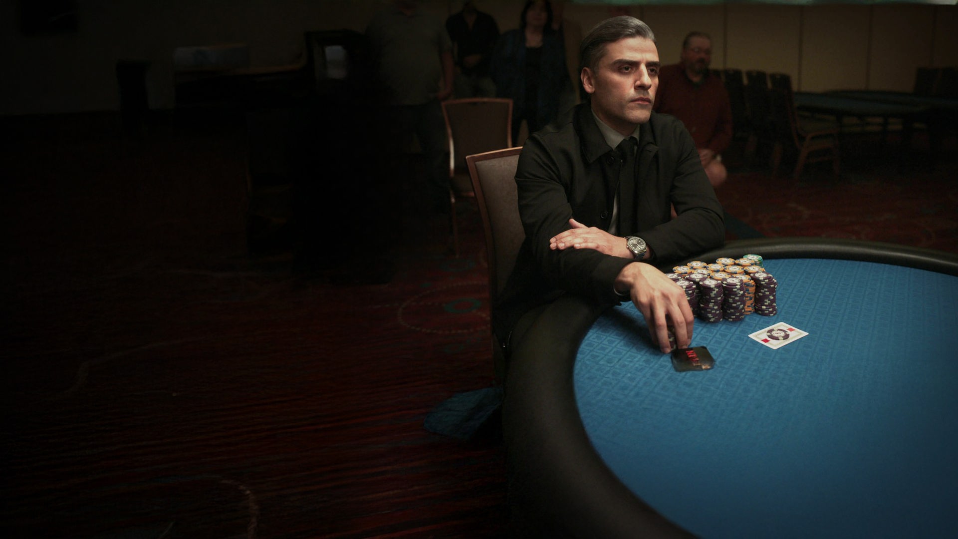 "The card counter" William Tell (Oscar Isaac) sitzt mit fokussiertem Blick in dunkler Kleidung an einem blauen Pokertisch, auf dem hohe Jetons-Stapel und Spielkarten liegen. Er hat die Arme verschränkt, trägt eine Armbanduhr und berührt mit einer Hand weitere Pokerchips vor sich.