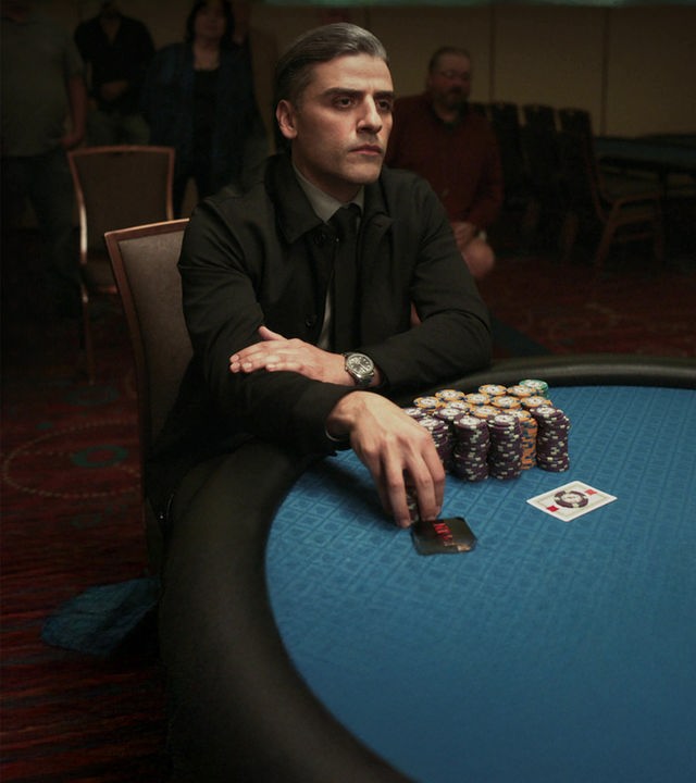 "The card counter" William Tell (Oscar Isaac) sitzt mit fokussiertem Blick in dunkler Kleidung an einem blauen Pokertisch, auf dem hohe Jetons-Stapel und Spielkarten liegen. Er hat die Arme verschränkt, trägt eine Armbanduhr und berührt mit einer Hand weitere Pokerchips vor sich.