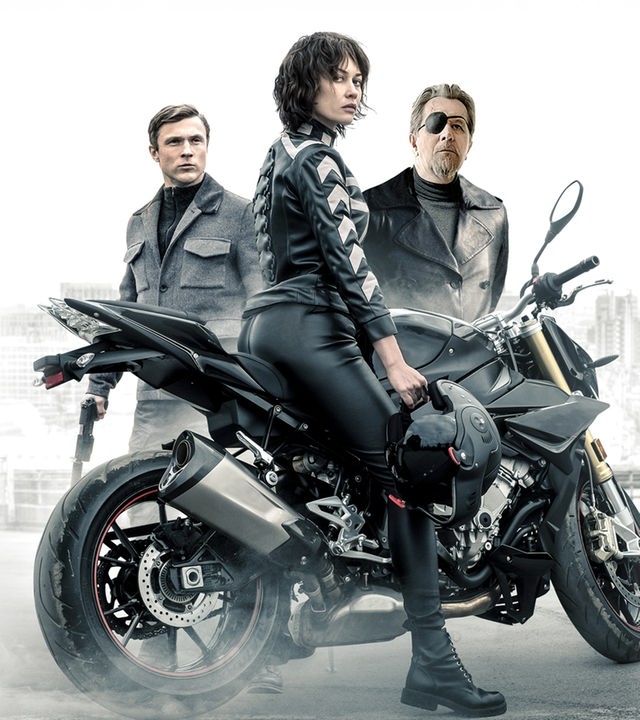 Der Kurier (Olga Kurylenko) sitzt auf einem Motorrad und sieht zur Kamera. Hinter dem Kurier sind Ezekiel Mannings (Gary Oldman) und Agent Bryant (William Moseley) zu sehen.
