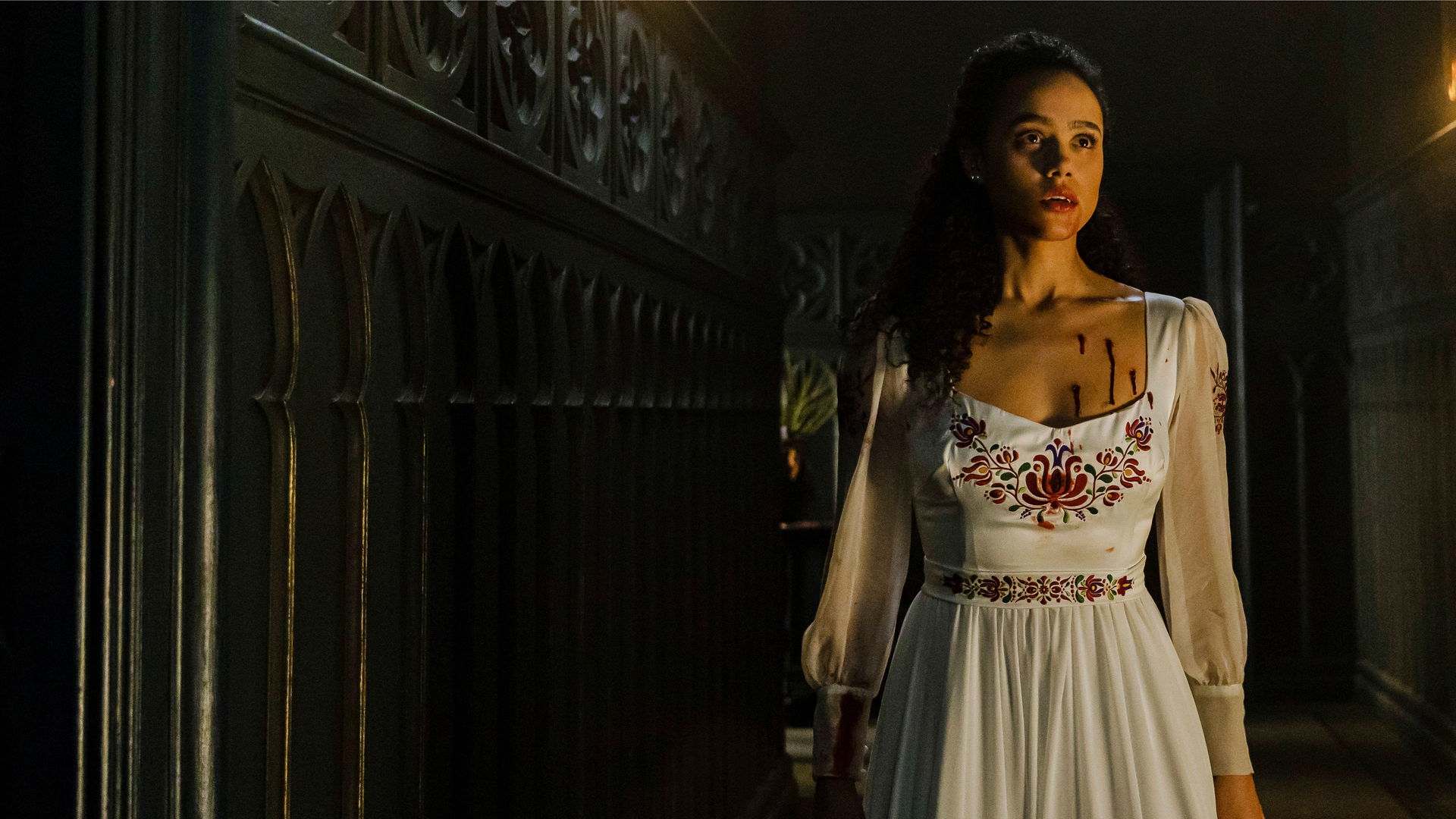 "The Invitation - Bis dass der Tod uns scheidet": Nahaufnahme: Mit geschocktem Blick steht Evie (Nathalie Emmanuel) im Brautkleid in einem dunklen Gang mit vertäfelten Holzwänden. An ihrer Brust sind mehrere blutige Einstichwunden zu sehen.