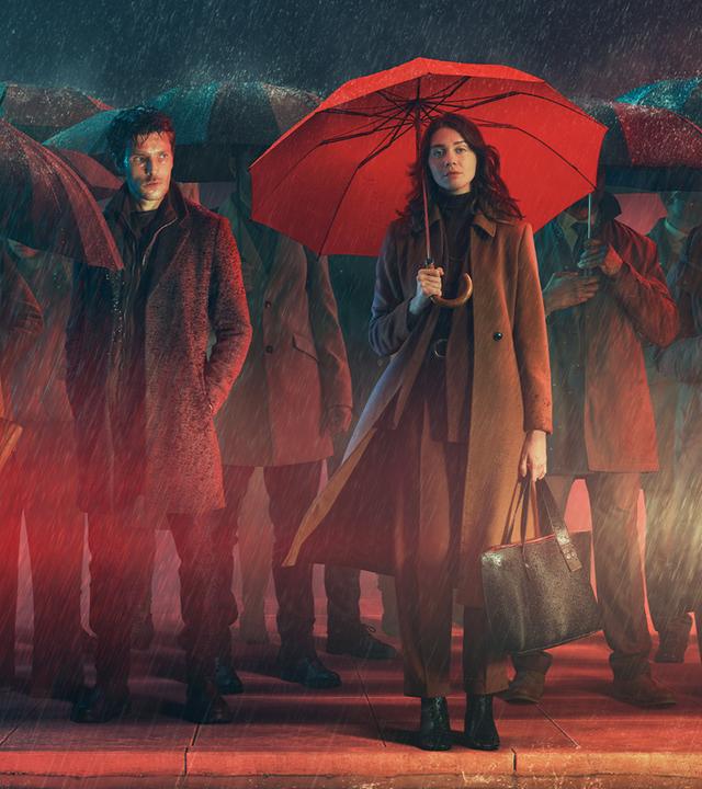 Stilisiertes Bild in Rot-Grau-Tönen, das eine Menschenmenge an einer Londoner Straße im Regen zeigt, vorne die beiden Hauptdarsteller, Ingrid Lewis (Emma Appleton) und John Webster (Colin Morgan). Sie hält einen roten Schirm über sich, er steht - als einziger ohne Schirm - daneben, die Hände in den Jackentaschen.