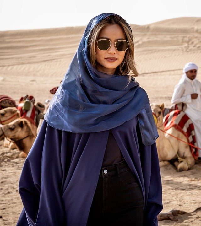 Hope (Hermione Corfield) trägt einen Kaftan und Sonnenbrille. Im Hintergrund seiht man einen Einheimischen und Dromedare.