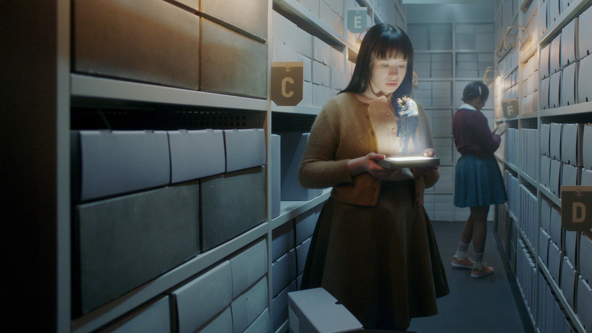 "The Ordinaries": Paula (Fine Sendel) steht im Gang eines Archivs und schaut auf ein Hologramm, welches sie in ihren Händen hält.  