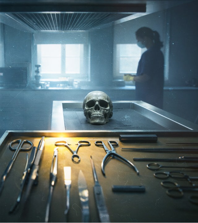 Ein dunkler Raum in der Gerichtsmedizin. Im Vordergrund sieht man einen Edelstahltisch mit verschiedenen Werkzeugen. Auf dem Seziertisch dahinter liegt ein menschlicher Schädel, der den Betrachter anschaut.