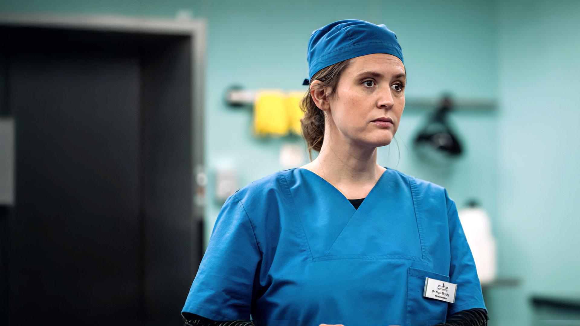 Mala Murphy (Emily Cox) befindet sich in der Pathologie und trägt blaue Schutzkleidung.