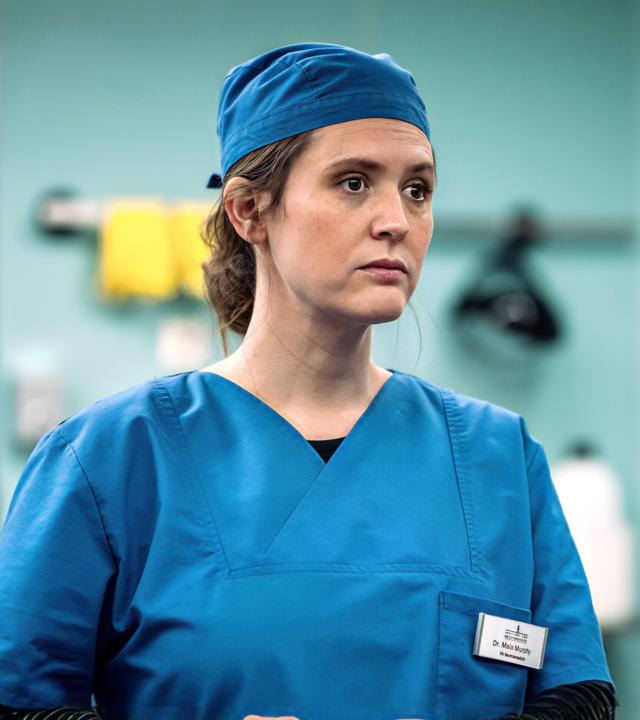 Mala Murphy (Emily Cox) befindet sich in der Pathologie und trägt blaue Schutzkleidung.