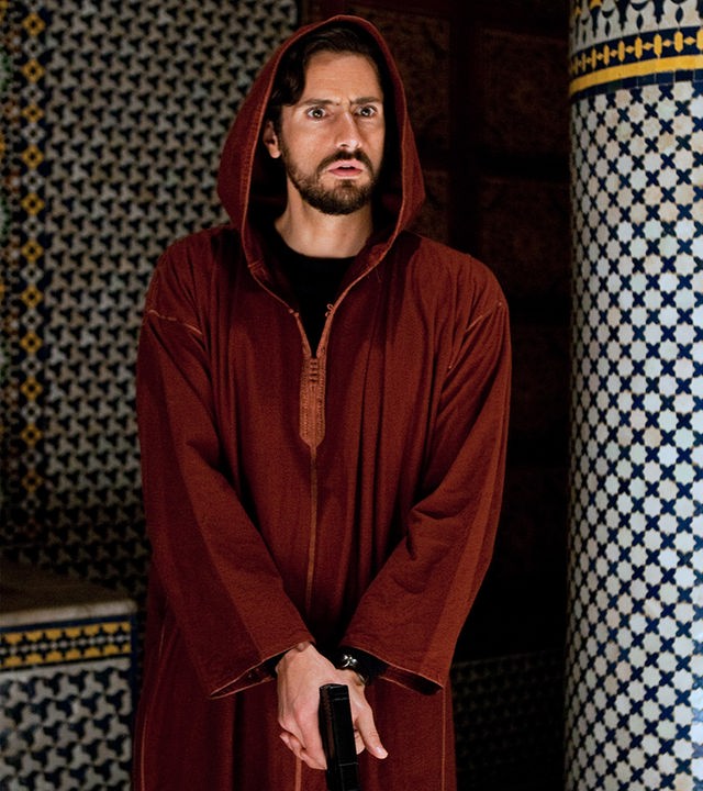 Kommissar Falcón (Juan Diego Botto) steht mit der Pistole in der Hand neben einer Säule und beobachtet entgeistert etwas.