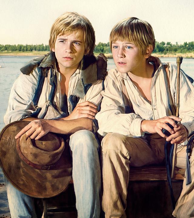 Tom Sawyer und Huckleberry Finn sitzen am Mississippi River. Im Hintergrund sieht man einen der typischen Raddampfer. 