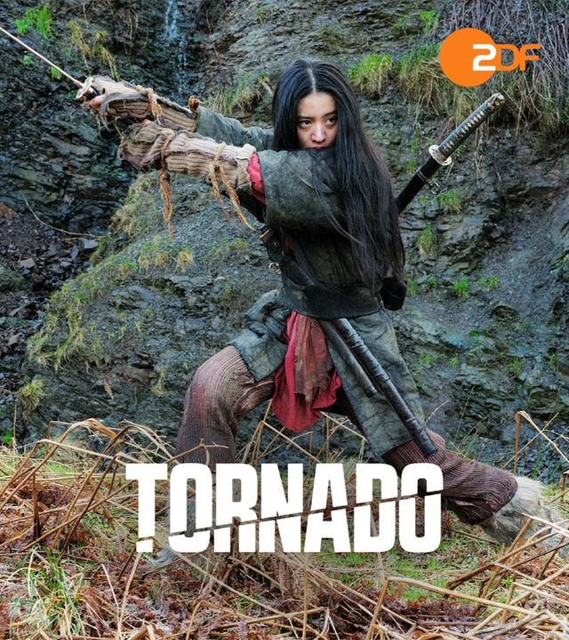"Tornado": Tagsüber auf einem Feld vor einem Felsen steht Tornado (Kôki,), eine junge japanische Frau im Teenageralter mit langen schwarzen Haaren und traditioneller Kleidung und schwingt ein Schwert vor sich her. Auf ihrem Rücken steckt ein weiteres Schwert.