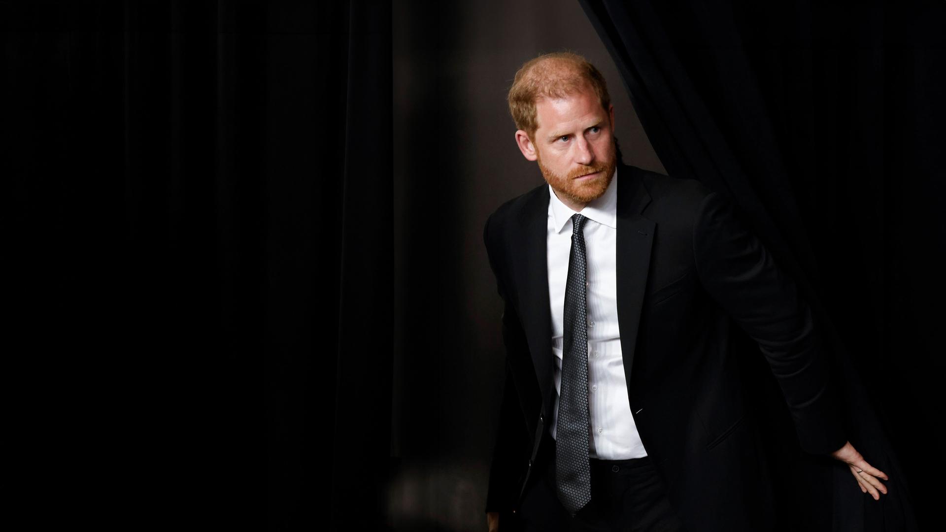 Prinz Harry schiebt einen schwarzen Vorhang zur Seite um durch den Spalt hindurchzugehen.