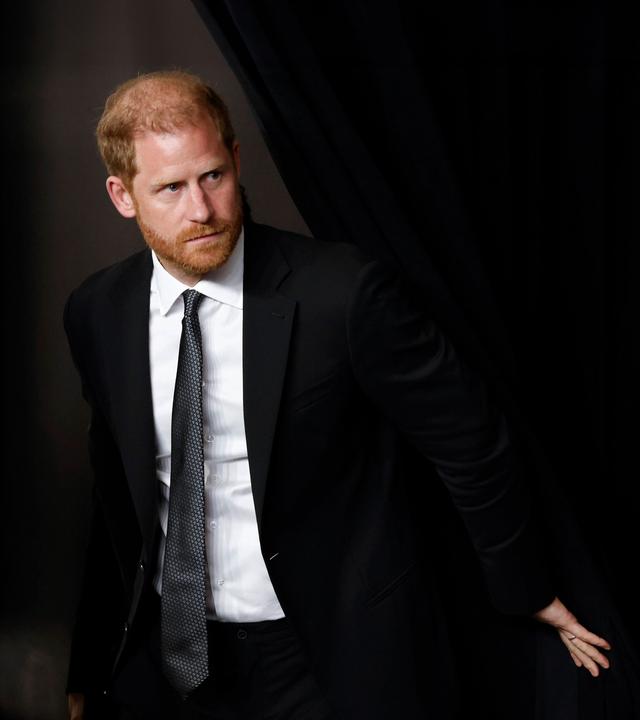 Prinz Harry schiebt einen schwarzen Vorhang zur Seite um durch den Spalt hindurchzugehen.