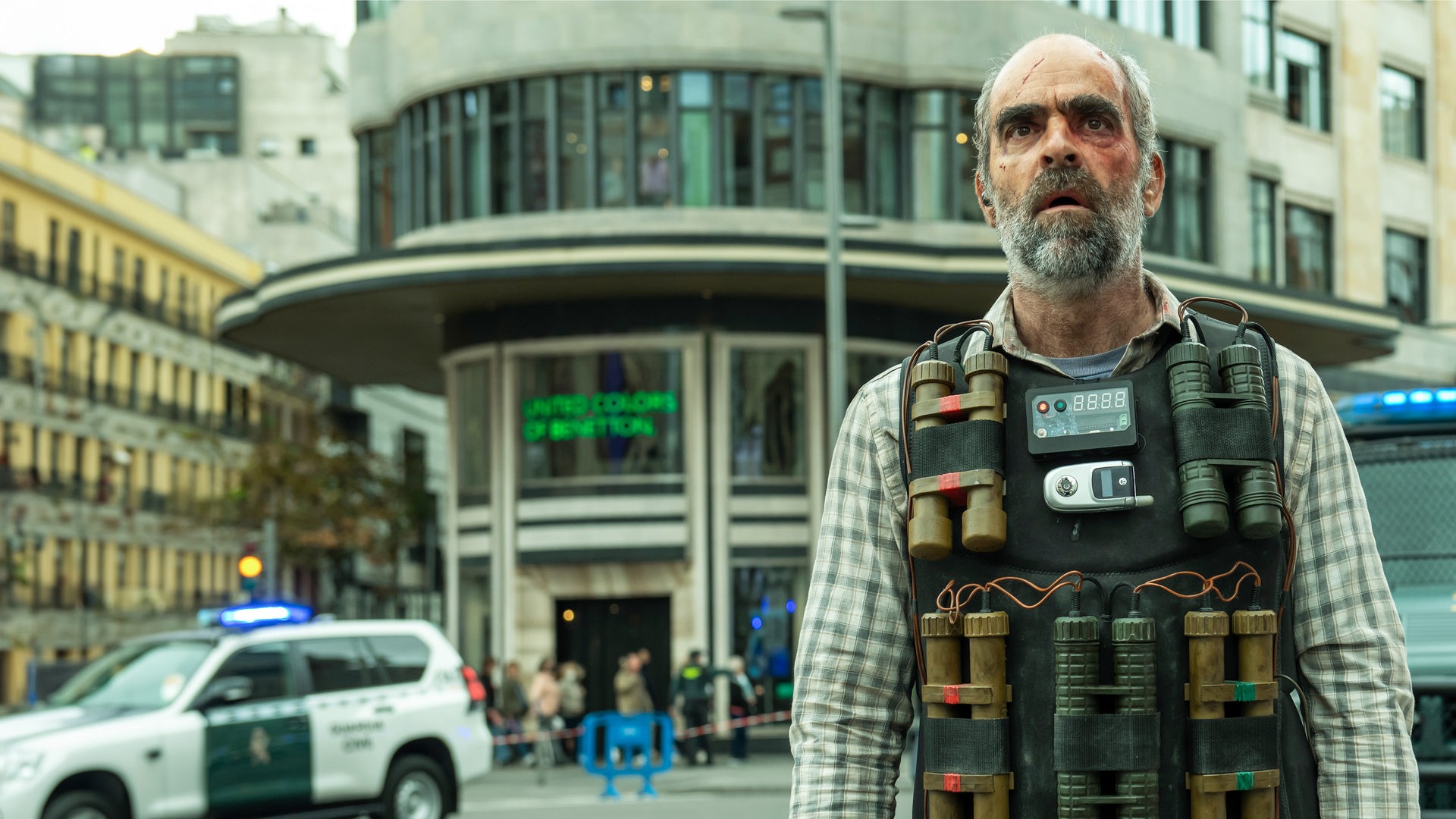 Halbnahe, leichte Untersicht - Santiago (Luis Tosar) mit Sprengstoffweste am Körper und fassungslosem Gesichtsausdruck auf abgesperrter, breiter Einkaufsstraße. Hinter Santiago sind mehrere Einsatzwägen der Polizei, sowie unscharf eine Gruppe von Schaulustigen zu erkennen.