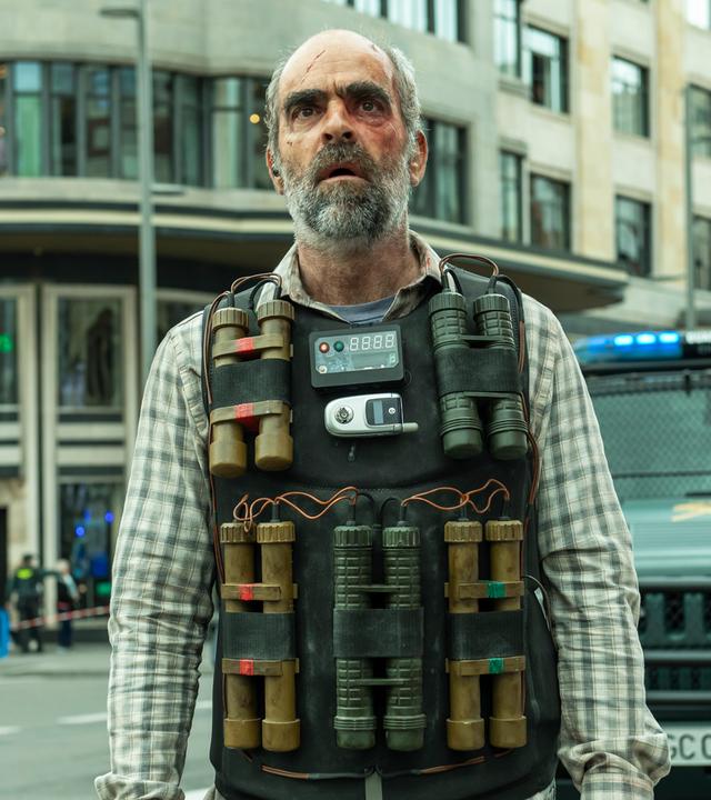 Halbnahe, leichte Untersicht - Santiago (Luis Tosar) mit Sprengstoffweste am Körper und fassungslosem Gesichtsausdruck auf abgesperrter, breiter Einkaufsstraße. Hinter Santiago sind mehrere Einsatzwägen der Polizei, sowie unscharf eine Gruppe von Schaulustigen zu erkennen.