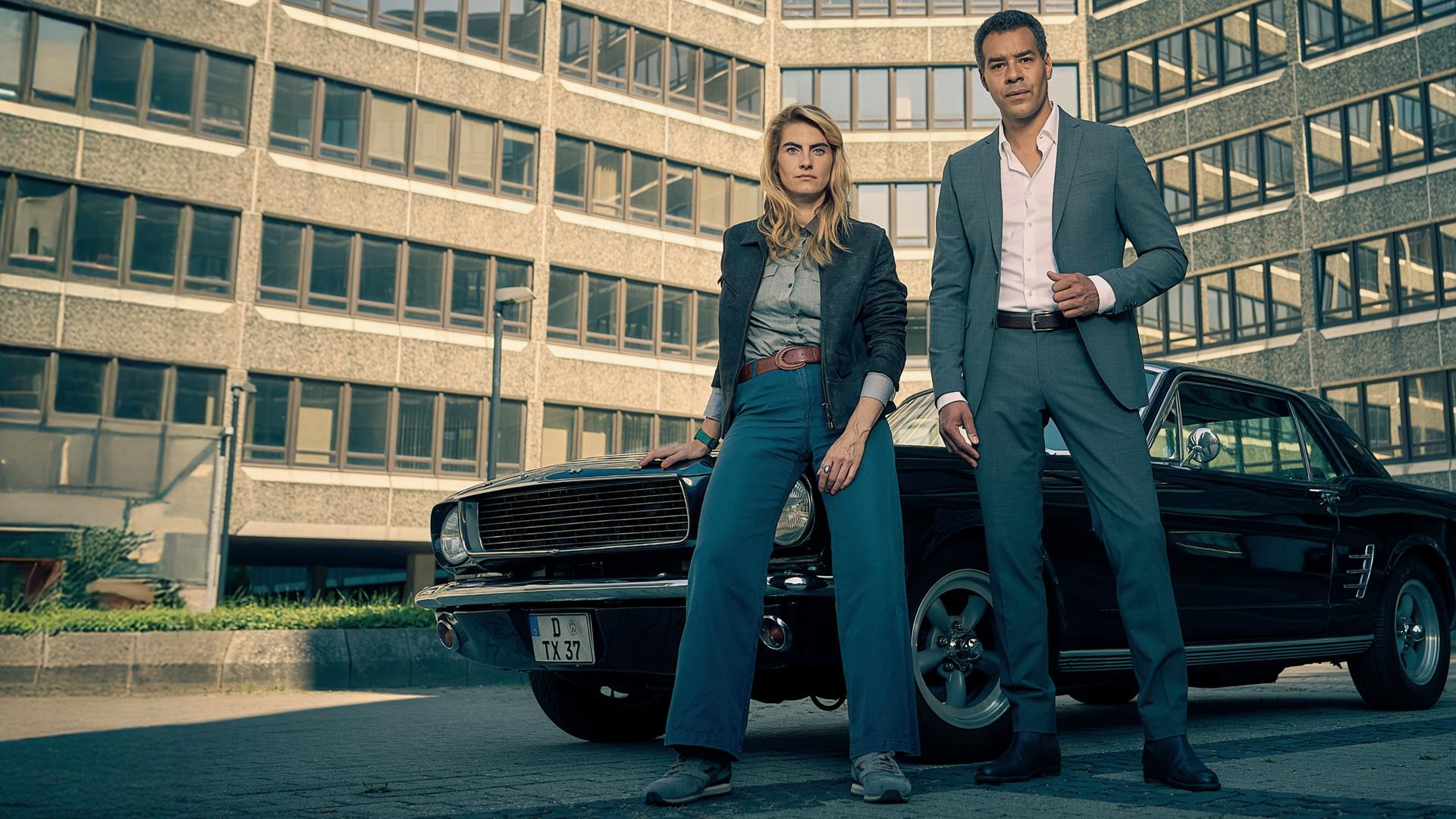 Clarissa Jakobs (Samia Chancrin) lehnt lässig an einem schwarzen Oldtimer. Neben ihr steht Joseph Kanjaa (Michael Klammer) in einem Anzug. Der gesamte Hintergrund wird von einem Bürogebäude ausgefüllt.