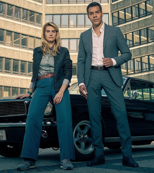 Clarissa Jakobs (Samia Chancrin) lehnt lässig an einem schwarzen Oldtimer. Neben ihr steht Joseph Kanjaa (Michael Klammer) in einem Anzug. Der gesamte Hintergrund wird von einem Bürogebäude ausgefüllt.