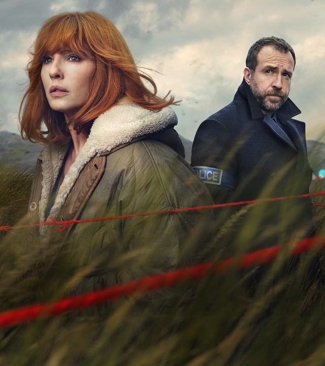 Under Salt Marsh: Die ehemalige Polizistin Jackie Ellis (Kelly Reilly) mit rotem Haar und wärmendem Mantel und der Polizist Eric Bull (Rafe Spall) in dunkler Uniform stehen inmitten eines windgepeitschten Kornfeldes unter einem stürmischen Himmel. Eine rote Schnur trennt den Tatort im Vordergrund ab.