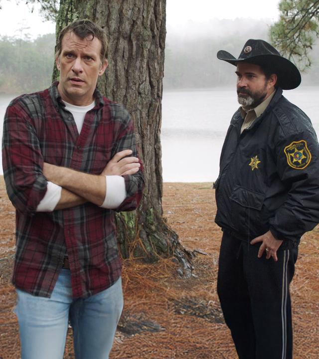  Paul (Thomas Jane) steht mit verschränkten Armen vor einem Baum. Rechts neben ihm steht Sheriff Baker (Jason Patric), im Hintergrund sind Wälder und der See zu sehen.