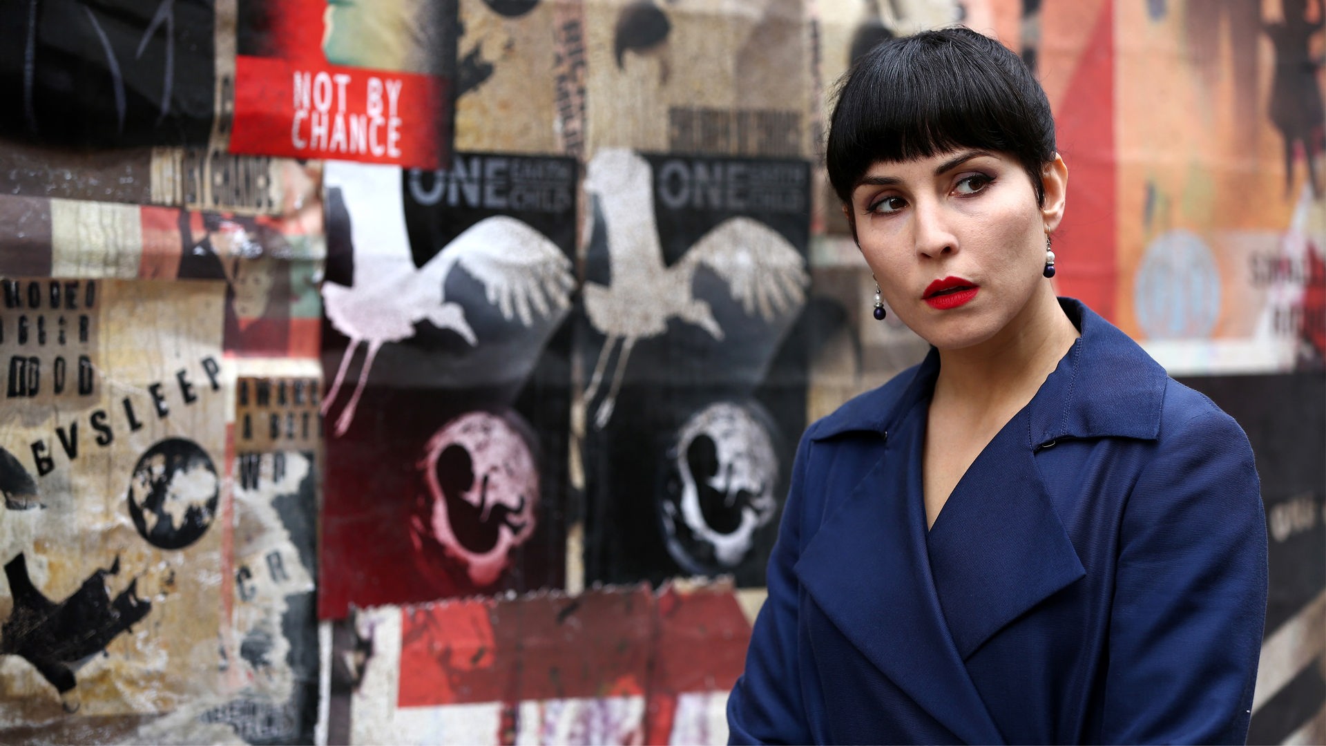  Monday (Noomi Rapace) steht vor einer plakatierten Mauer und sieht zur Seite.