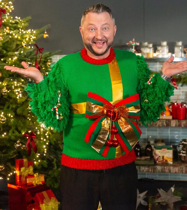 Sebastian Lege trägt einen Weihnachtspulli mit großer Schleife und steht vor einem Weihnachtsbaum. 