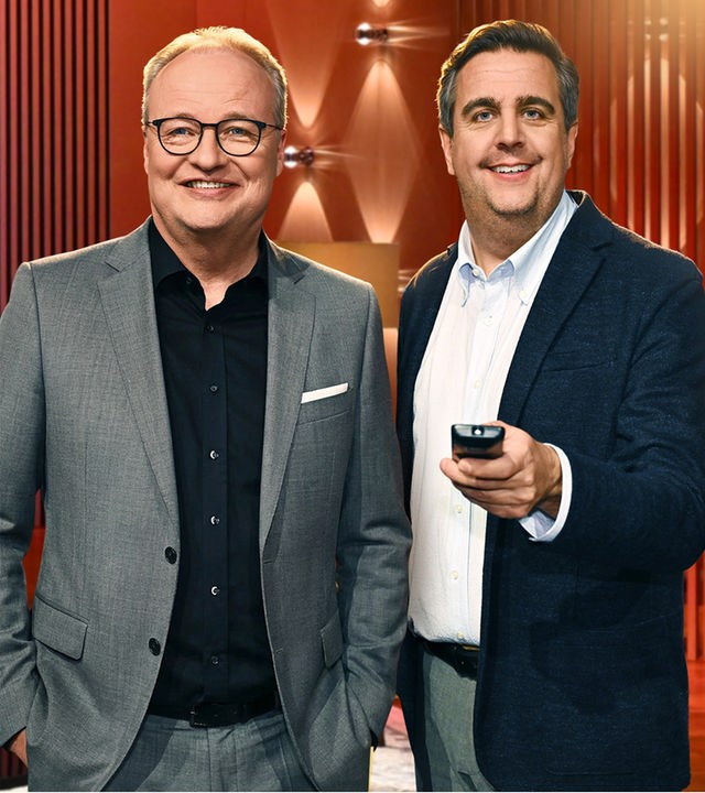 Oliver Welke und Bastian Pastewka, mit Fernbedienung in der Hand, posieren nahe beieinander für die Kamera im Set ihrer Show. Beide schauen direkt in die Kamera.