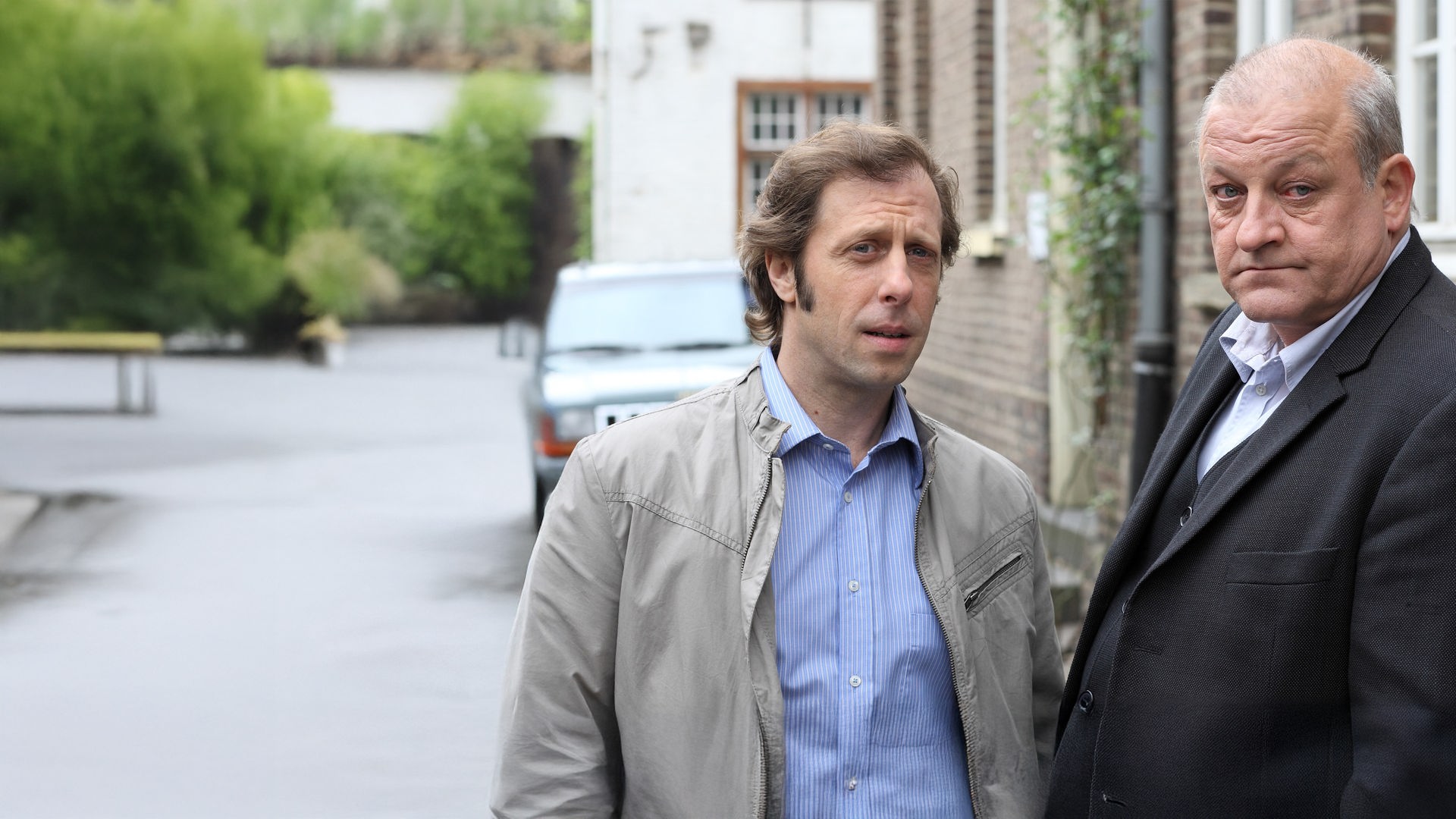 "Wilsberg - Aus Mangel an Beweisen": Außenaufnahme. Ekki (Oliver Korittke) und Wilsberg (Leonard Lansink) stehen nebeneinander vor einer Backstein-Hausfassade.