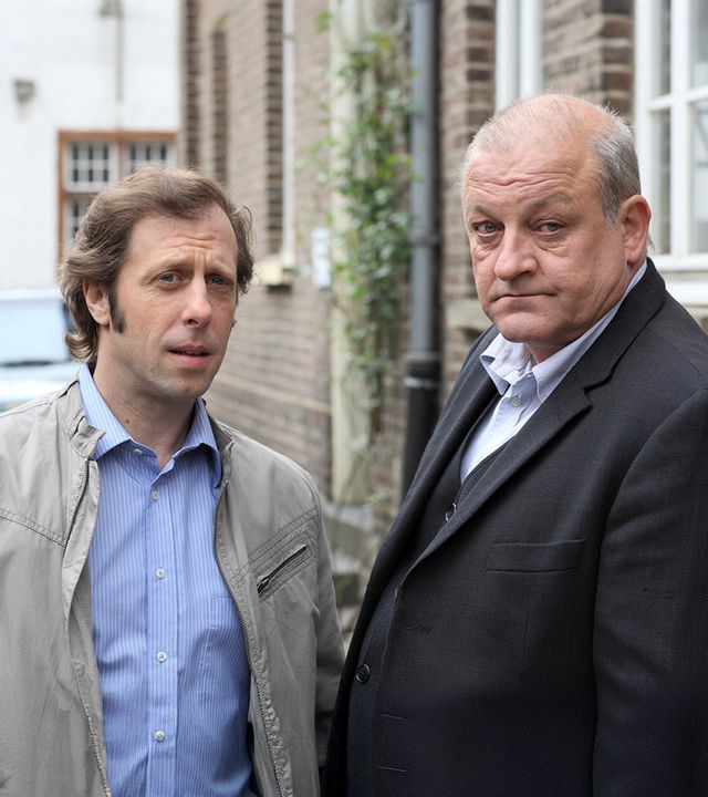 "Wilsberg - Aus Mangel an Beweisen": Außenaufnahme. Ekki (Oliver Korittke) und Wilsberg (Leonard Lansink) stehen nebeneinander vor einer Backstein-Hausfassade.