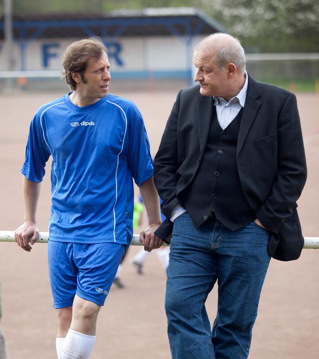 "Wilsberg - Aus Mangel an Beweisen": Ekki (Oliver Korittke) trägt Fußballkleidung und unterhält sich mit Georg Wilsberg (Leonard Lansink), an die Reling gelehnt, auf einem Sportplatz.