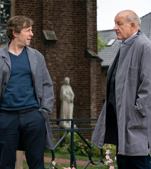 „Wilsberg – Einer von uns“: Vor einem Schloss stehen Wilsberg (Leonard Lansink) und Ekki (Oliver Korittke) und schauen sich an. Sie tragen Arbeitskittel und haben lässig die Hände in die Taschen gesteckt.