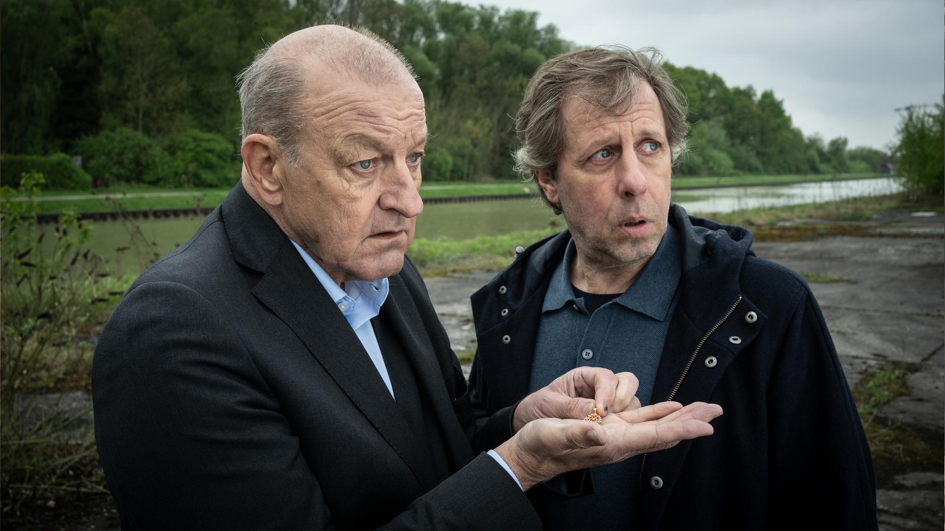 Wilsberg - Einfach weg": Wilsberg (Leonard Lansink) und Ekki (Oliver Korittke) stehen an einem Kanalufer. Wilsberg hält in seiner ausgestreckten Hand eine Goldkette.